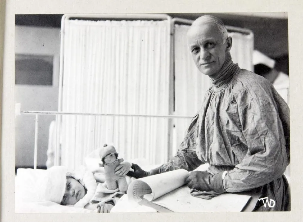 Black and white photograph with man (Cushing) dressed in surgical scrubs standing over a young child in a hospital bed. They are both staring at the camera.